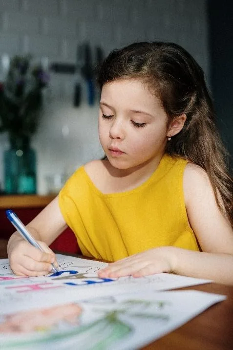 a little girl is sitting at a table drawing with a pen