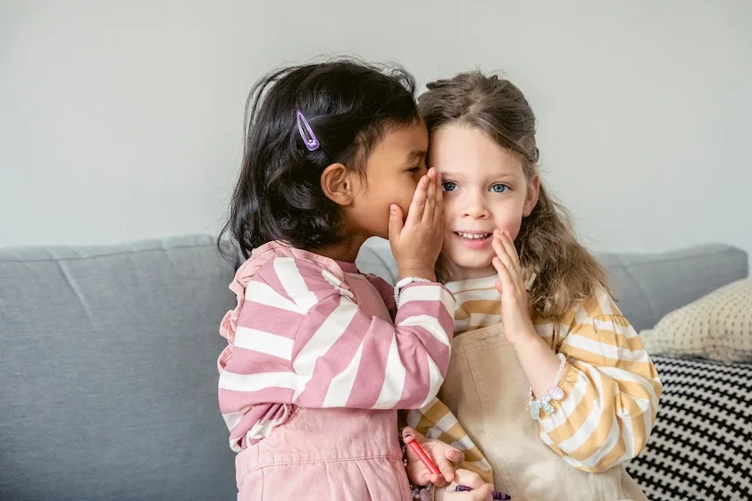two little girls whispering into each other 's ears
