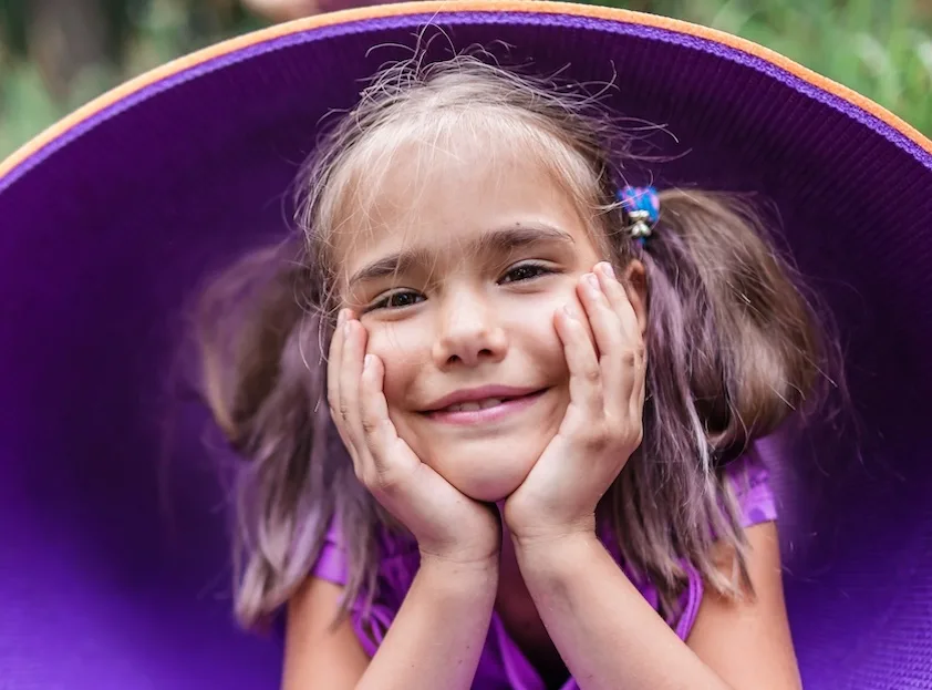 a little girl with purple hair is smiling with her hands on her face
