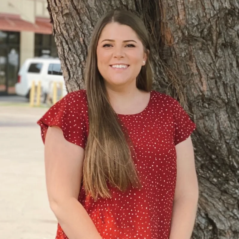 a woman in a red polka dot shirt smiles in front of a tree