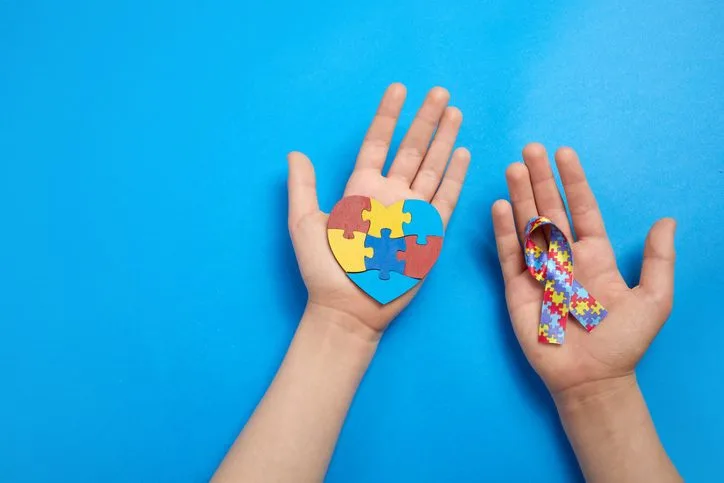 Adult and autistic boy hands holding puzzle heart shape and ribbon. Autism spectrum disorder family support concept. World Autism Awareness Day