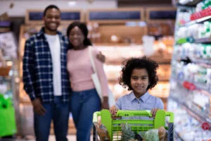 Happy little black girl with shopping trolley and her parents buying goods at modern supermarket