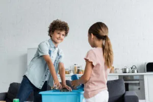 Smiling boy putting tin cans in box near blurred sister at home