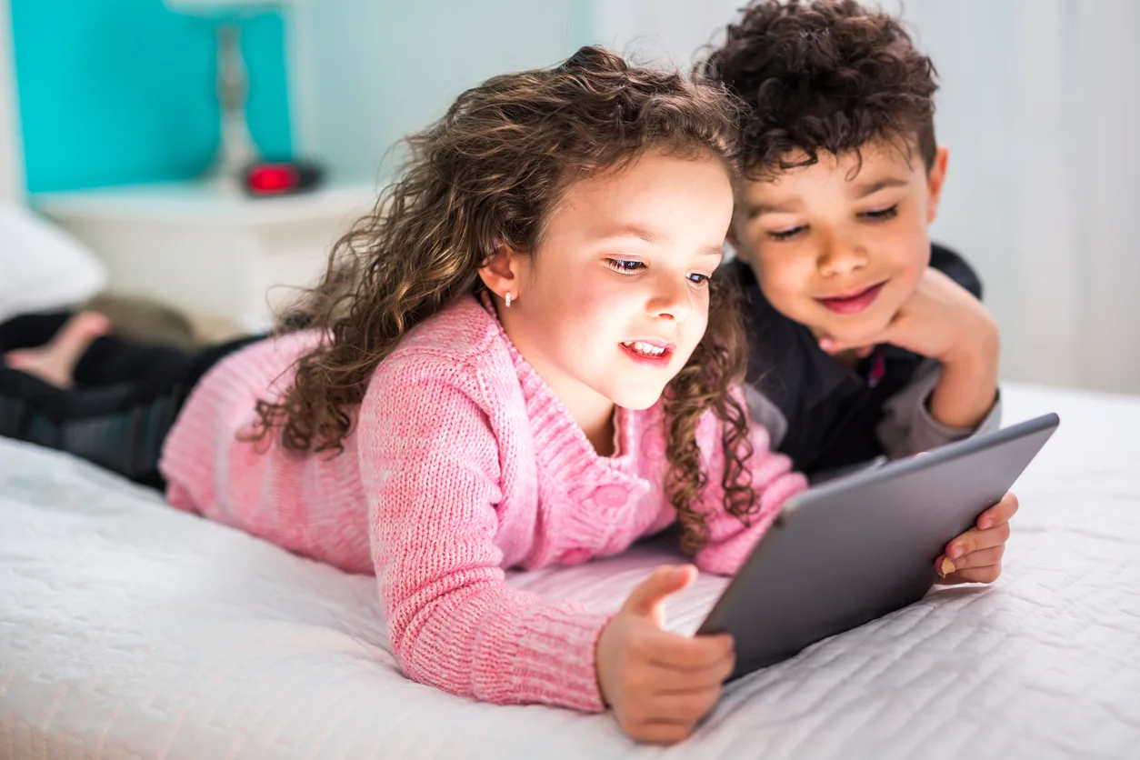 sibling playing on ipad laying on a bed