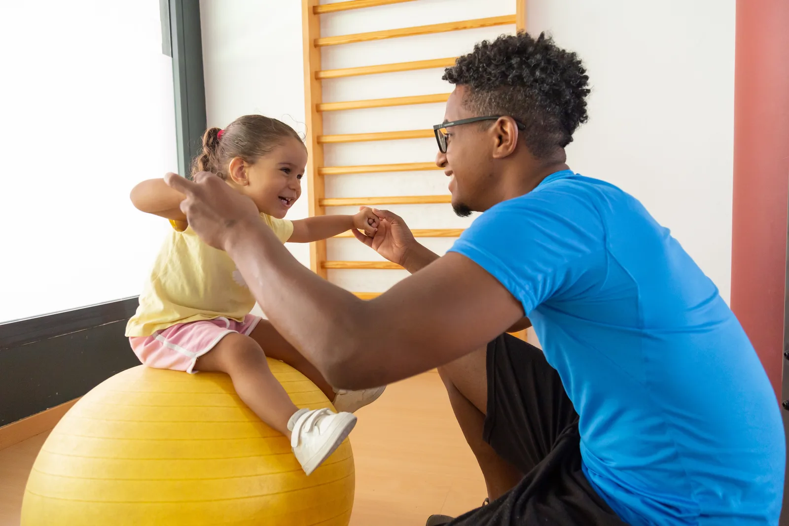 Side view of African American dad and mixed race child holding yellow fit ball in raised arms during fitness workout in gym