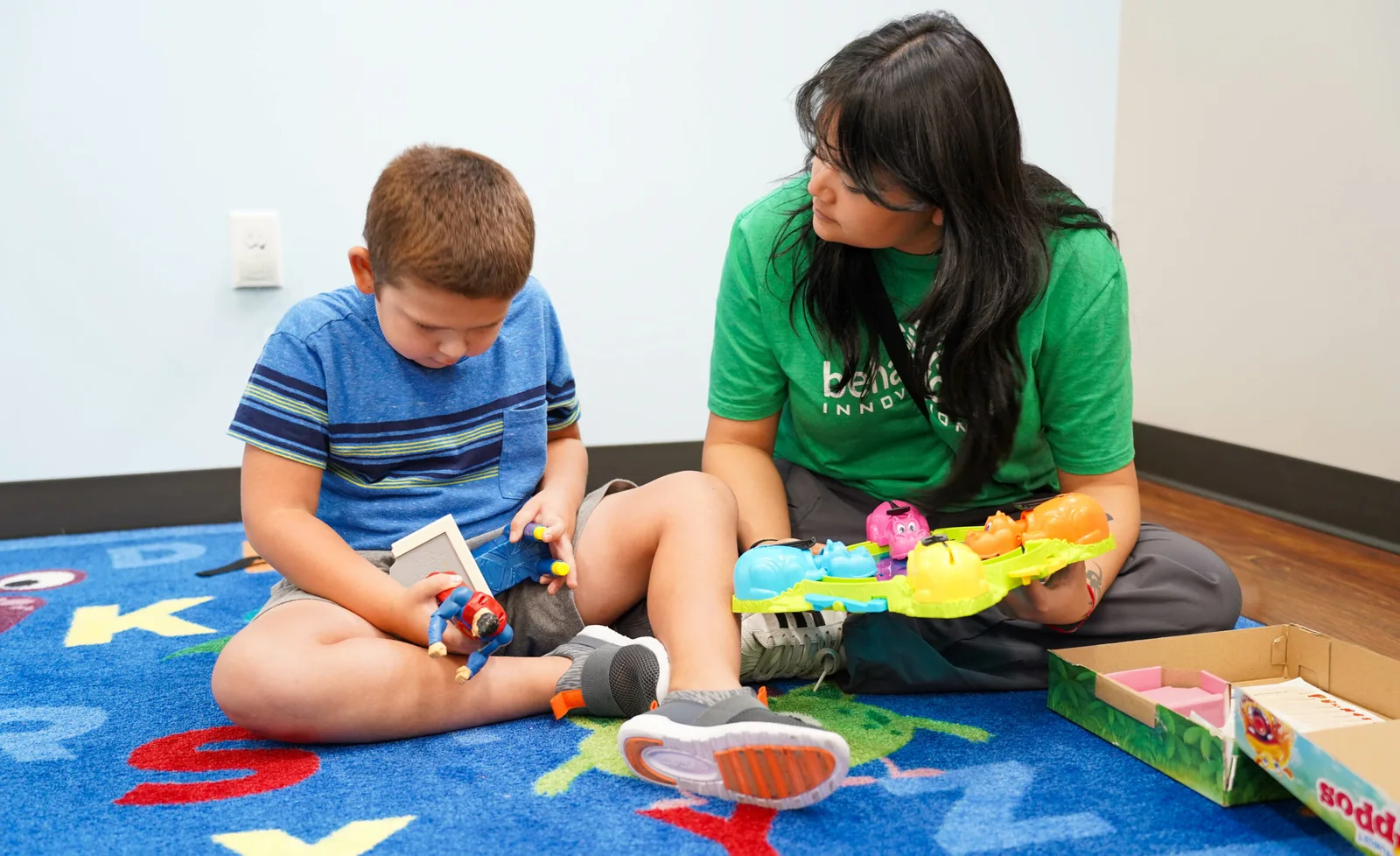 aba therapist in a green shirt is sitting on the floor with a young boy playing with toys