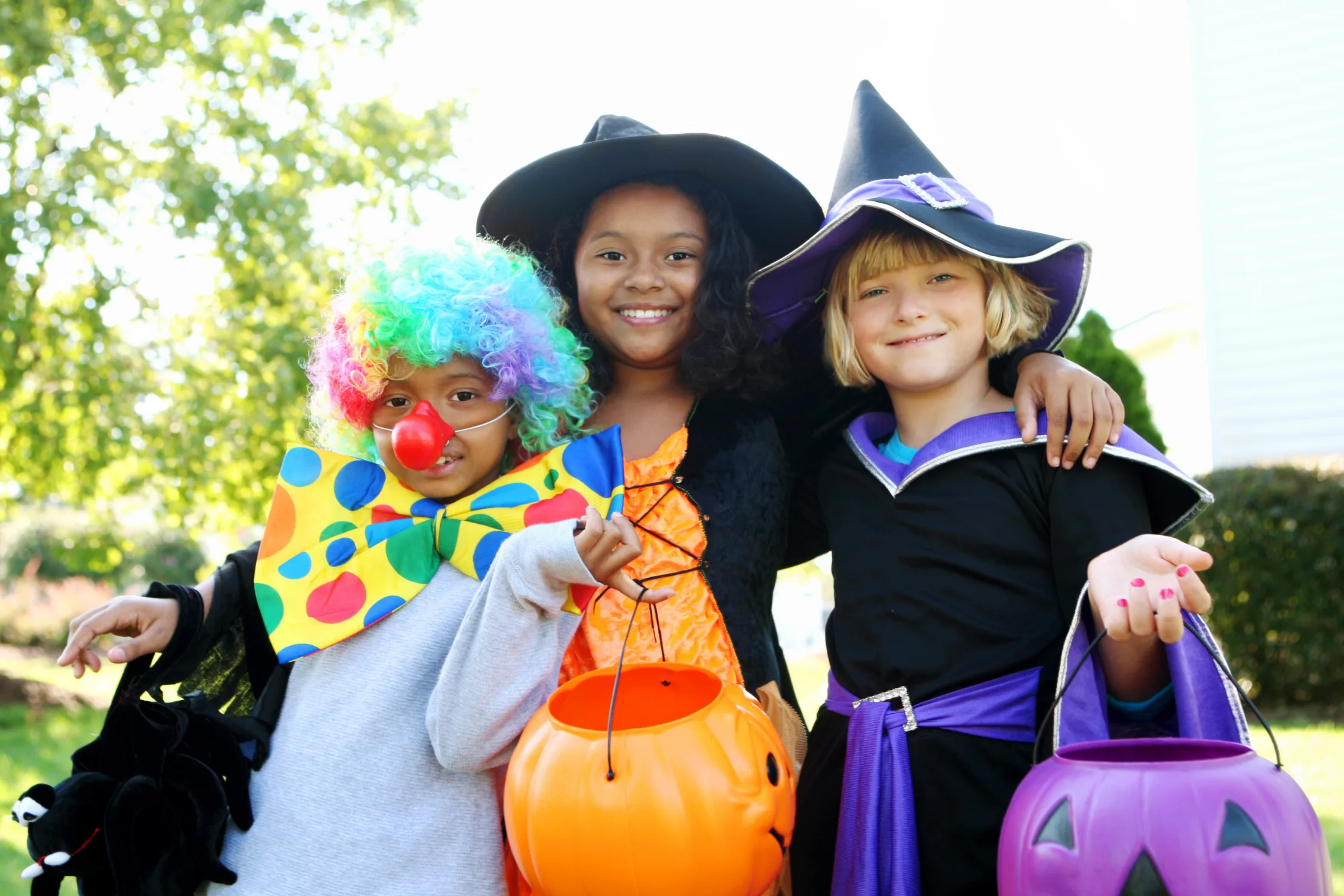Three children smile in Halloween costumes, including a clown and witches, holding trick-or-treat buckets outdoors.