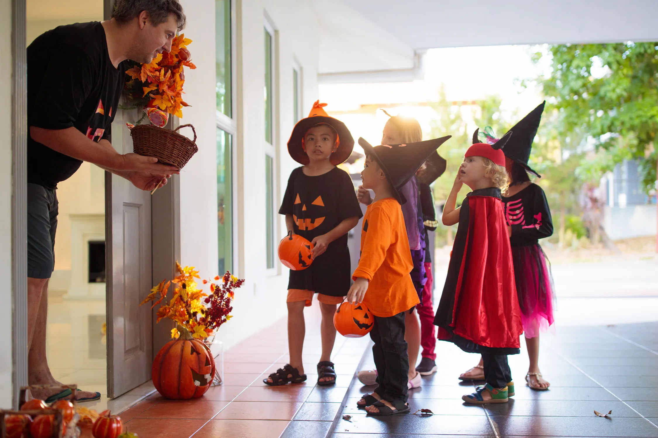 A homeowner offers a treat basket to children in Halloween costumes waiting on a decorated porch during trick-or-treating.